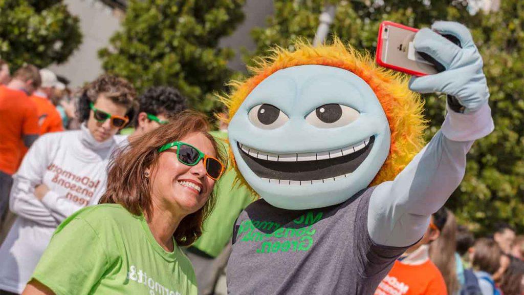 A staff member in orange and green sunglasses poses for a selfie with UTD mascot Temoc.